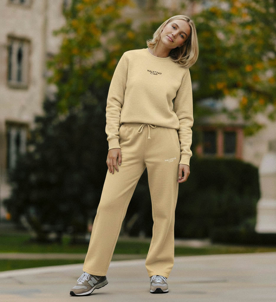 Woman wearing a beige sweatshirt and pants set outdoors with trees and a building in the background.