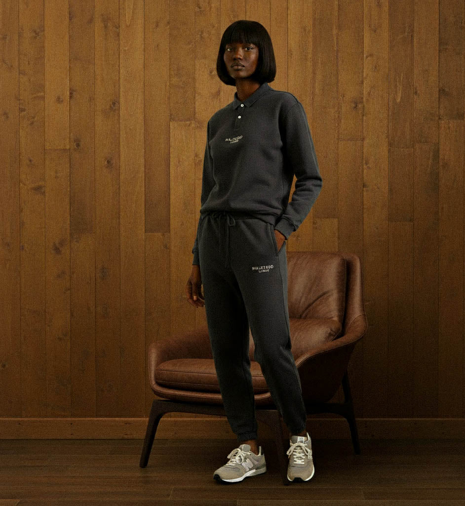 Person wearing a dark gray tracksuit standing against a wooden wall with a brown leather chair.
