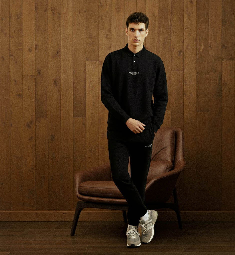 Man in a black outfit standing next to a brown chair against a wooden wall.