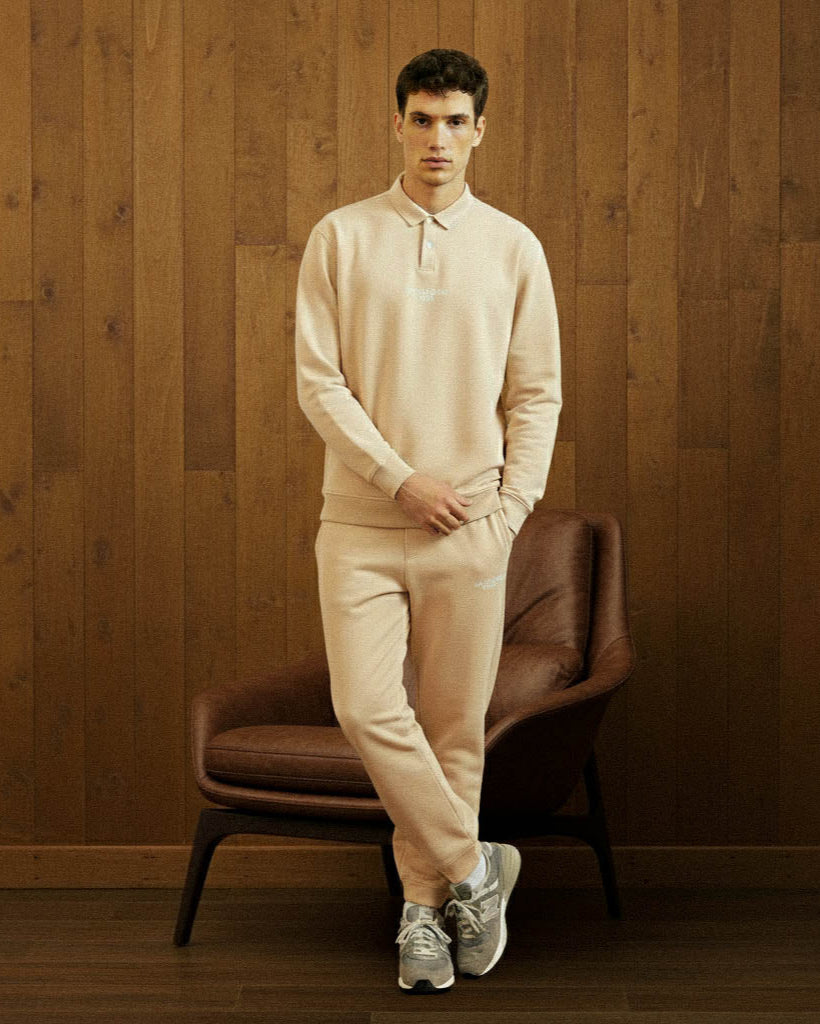 Man in beige outfit standing next to a brown chair against a wooden wall.