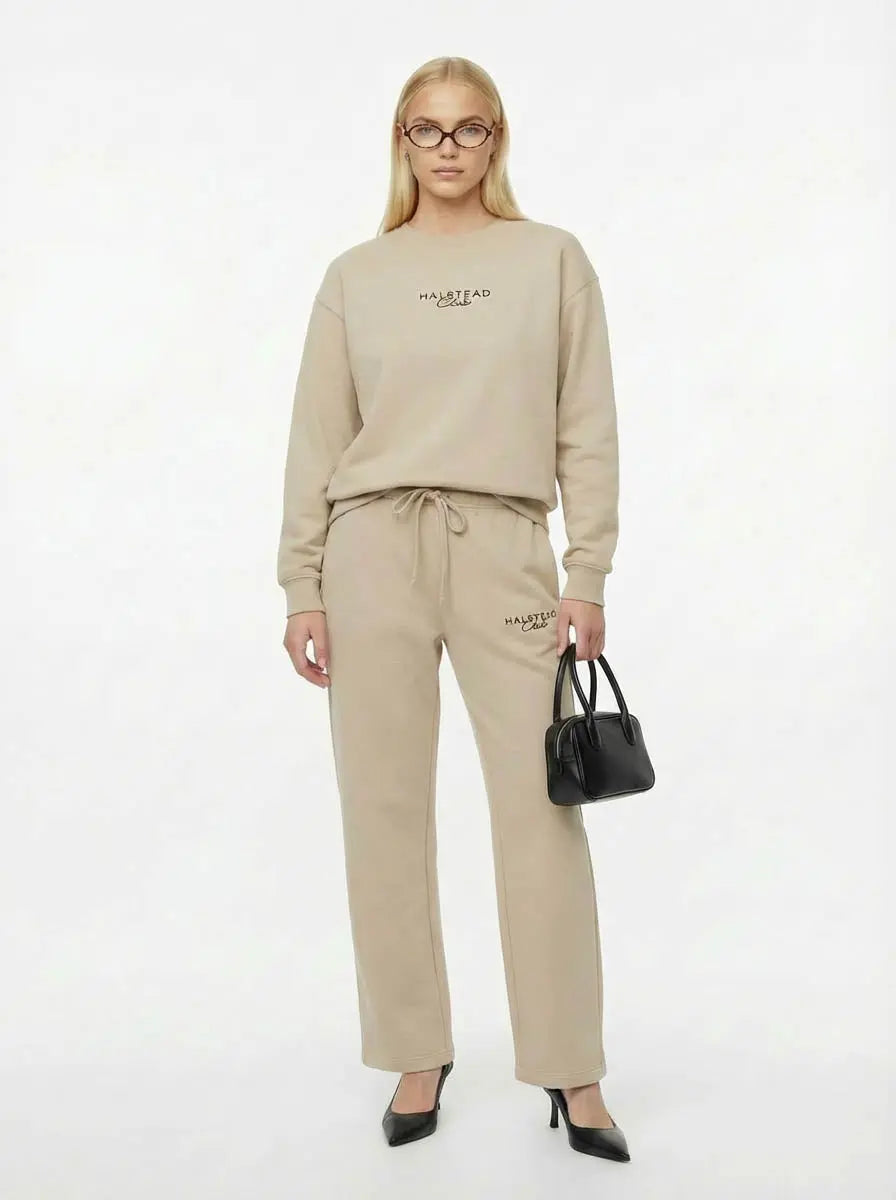 Woman wearing a beige sweatshirt and pants with 'HARPER' branding, holding a black handbag on a white background.