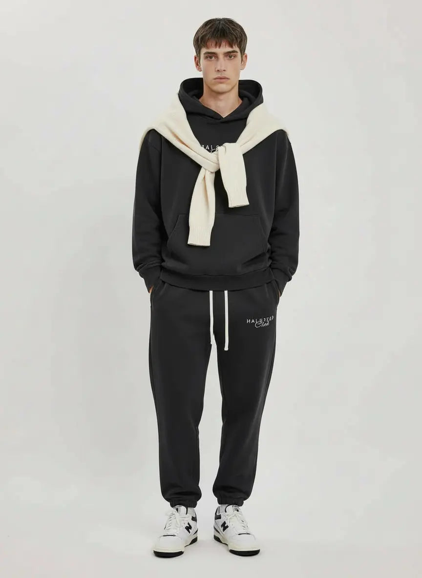 Person wearing a black hoodie and pants with a white scarf, standing against a plain background.