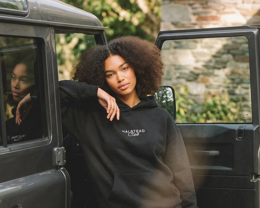 Person wearing a black hoodie with 'Halfstead Club' text, leaning against a vehicle.