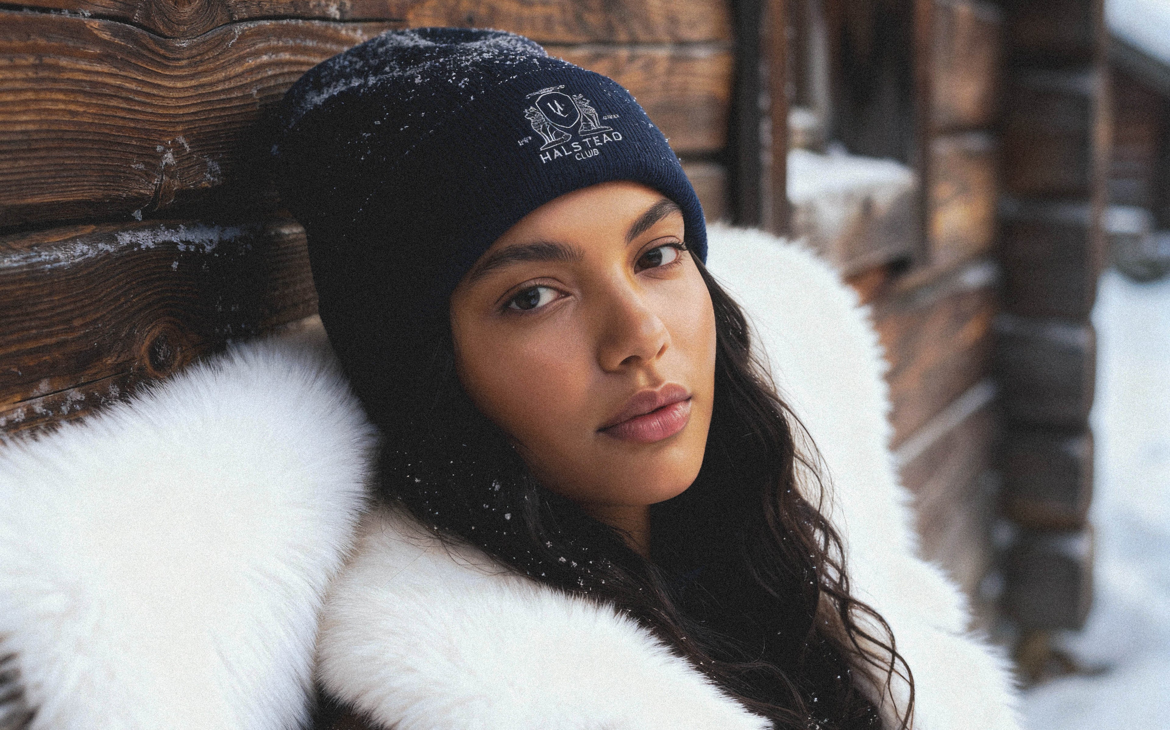 Woman wearing a black knit beanie with a logo and a white fur coat in a snowy setting.