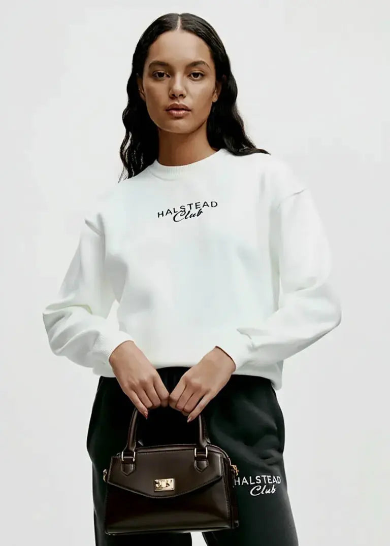 Woman wearing a white sweatshirt and black pants with 'Halstead Club' branding, holding a matching handbag.