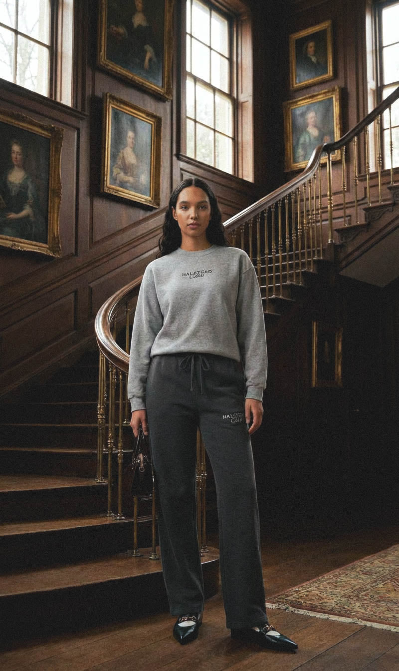 Woman in gray sweatshirt and pants standing in a grand staircase with wooden walls and framed portraits.