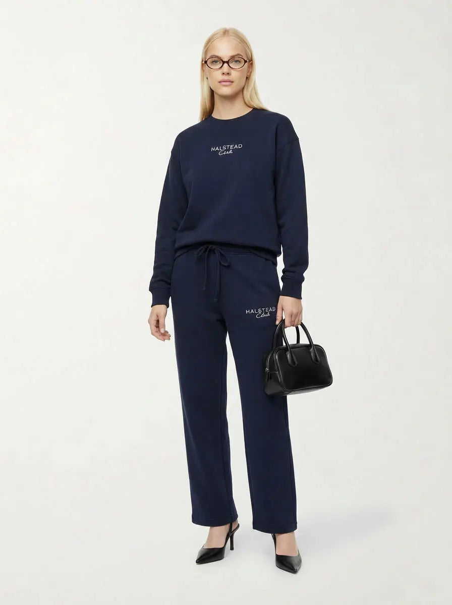 Woman wearing navy blue tracksuit with brand logo, holding a black handbag on a white background